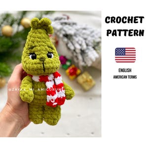 Könnte beinhalten: Eine grüne gehäkelte Figur mit schwarzen Augen und einem rot-weiß gestreiften Schal. Die Figur hält im Hintergrund einen Weihnachtsbaum. Der Text "CROCHET PATTERN" wird in Schwarz zusammen mit der amerikanischen Flagge und den Worten "ENGLISH AMERICAN TERMS" angezeigt.