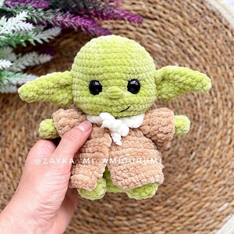 Op de afbeelding: Een handgemaakte gehaakte pop in groen en bruin, die lijkt op een personage uit een populaire sciencefictionfranchise. De pop heeft grote oren, zwarte ogen en draagt een bruine jurk met een witte kraag. De tekst "@ZAYKA MI AMIGURUMI" is zichtbaar.