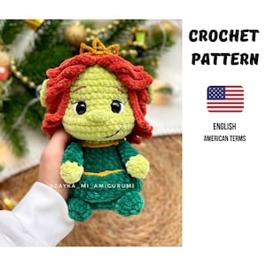 Puede incluir: Una muñeca de ganchillo hecha a mano con una cara verde, pelo rojo y una corona dorada. La muñeca lleva un vestido verde con detalles amarillos. La imagen incluye el texto "CROCHET PATTERN" y "ENGLISH AMERICAN TERMS" con la bandera americana.