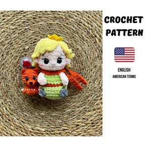 Puede incluir: Muñeca amigurumi de ganchillo de un personaje con pelo amarillo, una corona dorada y una bufanda naranja, sosteniendo un pequeño oso rojo. La muñeca lleva un atuendo verde y naranja. El texto "CROCHET PATTERN" y "ENGLISH AMERICAN TERMS" también es visible.