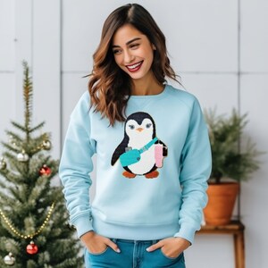 May include: A light blue sweatshirt with a penguin graphic. The penguin is wearing a blue and green backpack and holding a pink drink.