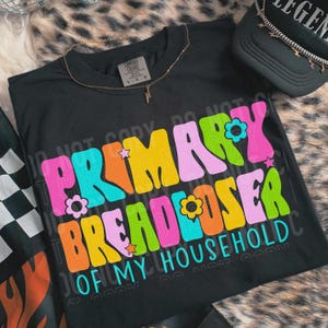 May include: A black t-shirt featuring the text "PRIMARY BREADLOSER OF MY HOUSEHOLD" in a multi-colored, wavy, retro-style font. The design includes small flower and star icons in pink, orange, yellow, lime green, and turquoise.