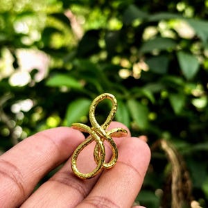 Puede incluir: Un anillo de tono dorado que presenta un diseño abstracto de bucles continuos con una superficie texturizada y martillada. La banda de metal forma una forma intrincada y multicapa que se curva y se superpone, creando un aspecto orgánico.