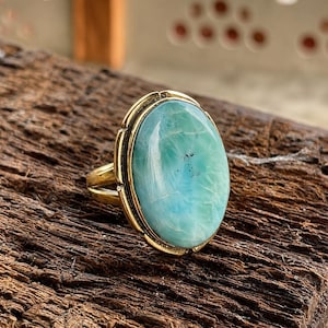 May include: A gold ring featuring a large, oval-shaped, light blue Larimar gemstone with natural white veining and small dark inclusions. The stone is set in a smooth gold bezel with a decorative scalloped edge and a simple, polished gold band.