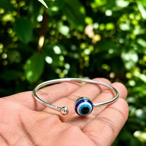Könnte beinhalten: Ein silberfarbenes Armband in offener Reifenform mit einem runden Nazar-Amulett, das konzentrische Ringe in Dunkelblau, Hellblau und Weiß aufweist, sowie einer kleinen polierten silbernen Kugel am anderen Ende des verstellbaren Metallbands.