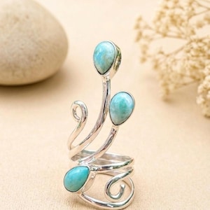 May include: A silver ring featuring three teardrop-shaped light blue stones set in polished metal. The band consists of swirling, curved silver wire elements that extend upward, holding each stone in a distinct position along the metal frame.
