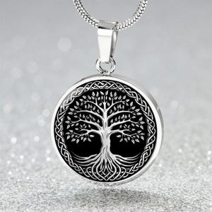 Puede incluir: Collar con colgante plateado con un colgante circular que muestra un diseño detallado del árbol de la vida. El árbol es blanco sobre un fondo negro, enmarcado por un borde de nudos celtas. El collar tiene una cadena fina.