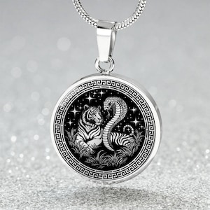 Puede incluir: Collar con colgante plateado. El colgante redondo presenta una ilustración en blanco y negro de un tigre y una cobra, sobre un fondo estrellado. El colgante tiene un borde decorativo y cuelga de una cadena plateada.
