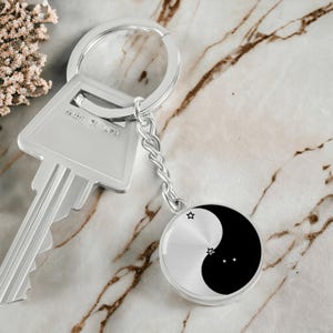 May include: A silver key chain with a black and white yin yang charm featuring a small constellation of stars. The charm is attached to a silver chain and a silver key ring.