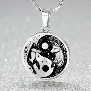 May include: A silver necklace with a round pendant featuring a yin yang symbol with two foxes on either side. The foxes are black and white and are facing each other. The pendant is surrounded by a black and white background with stars and flowers.