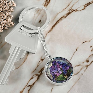 May include: A silver key chain with a round charm featuring a stained glass design of purple flowers. The charm is attached to a silver chain and a silver key ring.