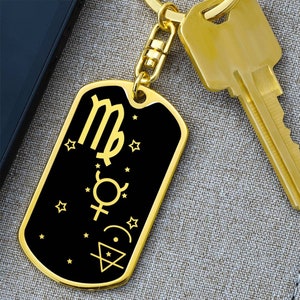May include: A gold-colored dog tag keychain with a black background. The tag features the Virgo zodiac symbol, other astrological symbols, and stars in gold. A gold key and a portion of a black object are in the background.