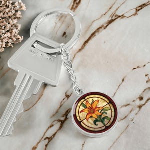 May include: Silver key chain with a round charm featuring a stained glass style image of an orange lily flower.
