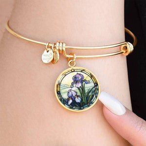 Pode incluir: Pulseira de ouro com um berloque redondo que apresenta uma imagem de estilo vitral de íris roxas. O berloque está preso a uma pulseira de bracelete de ouro com um fecho que diz "HAND MADE".