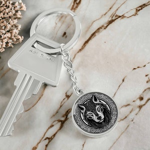 May include: A silver key chain with a round, silver charm featuring a black and white wolf head design with Celtic knotwork. The charm is attached to a silver chain and a silver key ring.
