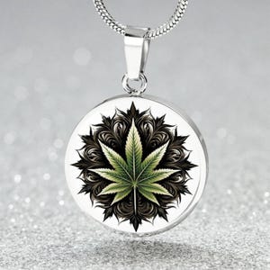 May include: A silver necklace with a round pendant featuring a green marijuana leaf with black and white details on a white background.