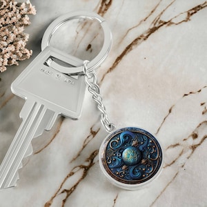 May include: A silver keychain with a round pendant featuring a celestial design in shades of blue, gold, and teal. The keychain includes a key and a decorative pendant with a detailed cosmic pattern.