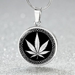 May include: A silver necklace with a round pendant featuring a black background and a white marijuana leaf design. The pendant has a decorative border.