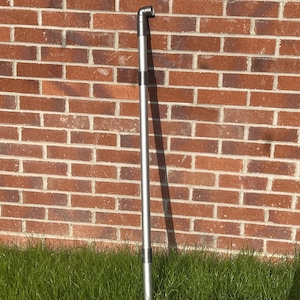 May include: A silver telescopic pole with a black plastic elbow joint at the top. The pole is standing upright against a brick wall and is partially obscured by green grass.