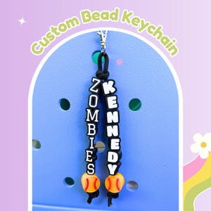 May include: Custom bead keychain with the names "ZOMBIES" and "KENNEDY" spelled out in black and white beads. The keychain includes two orange and white baseball charms and is attached to a silver clasp. The text "Custom Bead Keychain" is in the background.