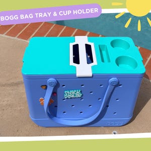 May include: A blue and teal Bogg Bag with a tray and cup holder. The bag has a handle and the text "Snack Dealer". The top of the bag has a teal tray with two cup holders and a slot. The image also includes the text "Bogg Bag Tray & Cup Holder".