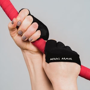 Puede incluir: Un par de guantes negros sin dedos con el texto "AERIAL ARMOR" en el dorso de la mano. Los guantes son usados por una persona que sostiene una barra roja.