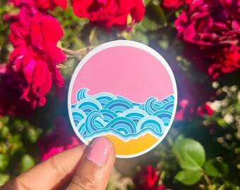Puede incluir: Una pegatina redonda con un cielo rosa, olas azules y una playa amarilla. La pegatina tiene un borde blanco. Se sostiene frente a un fondo de rosas rojas.
