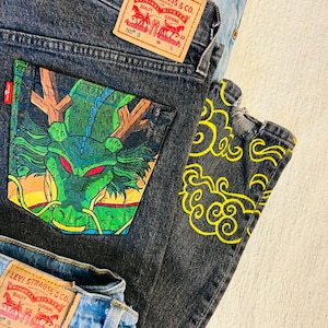 Puede incluir: Primer plano de tres pares de vaqueros Levi Strauss & Co. 501. Un par es negro con un diseño de dragón verde y amarillo en el bolsillo trasero. Otro par es azul claro. El tercer par es negro con diseños de nubes amarillas.