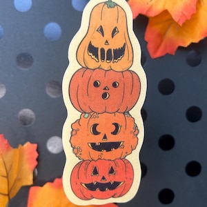 Puede incluir: Una pegatina con una pila de cuatro calabazas naranjas con caras talladas. Las calabazas son de diferentes tonos de naranja y tienen diferentes expresiones. La pegatina tiene un borde color crema y está sobre un fondo oscuro con lunares.