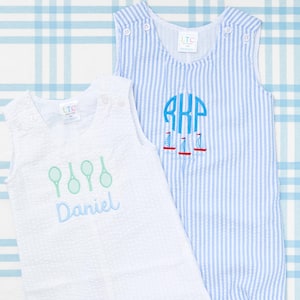 May include: Two white sleeveless baby rompers with blue and white stripes. One romper has a green tennis racket design and the name "Daniel" embroidered on it. The other romper has a blue monogram "RKP" and a red sailboat design.