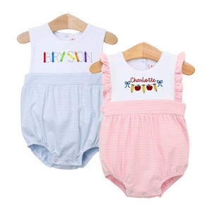 Könnte beinhalten: Zwei Baby-Strampler an Holzbügeln. Einer ist hellblau mit weißem Oberteil und dem Namen "Bryson" mit bunten Buntstiften bestickt. Der andere ist rosa gestreift mit weißem Oberteil und dem Namen "Charlotte" mit Bleistift- und Apfelmotiven.