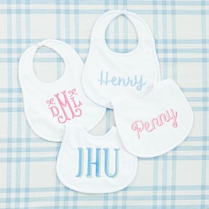 May include: Four white baby bibs with different embroidered designs. The bibs have a scalloped edge and are made of soft, absorbent fabric. The designs include a pink monogram "DML", a blue name "Henry", a pink name "Penny", and a blue and green name "JHU".