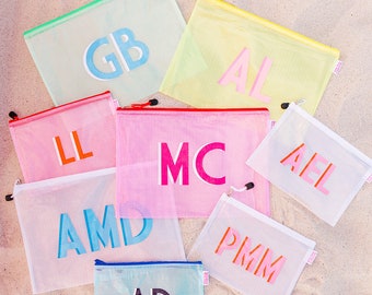 Puede incluir: Una colección de ocho bolsas de malla con cremallera de colores con diferentes iniciales impresas en ellas. Las bolsas están dispuestas sobre una superficie arenosa.