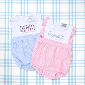 Puede incluir: Dos peleles de bebé, uno azul y otro rosa, con la parte superior blanca. El pelele azul tiene el nombre "HENRY" bordado en rojo y azul. El pelele rosa tiene el nombre "Colette" bordado en verde y rosa.