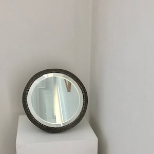 Puede incluir: Un espejo redondo, adornado, con marco plateado y borde con patrón en blanco y negro. El espejo está sobre un pedestal blanco.