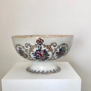 May include: A white ceramic bowl with a pedestal base, decorated with floral and foliate patterns in red, blue, and gold. The rim has a gold trim. The bowl is set against a plain white background.
