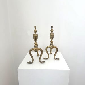 Op de afbeelding: Een paar bijpassende koperen haardbokken met een verticale centrale paal met een decoratieve knop aan de bovenkant en drie gebogen poten die eindigen in kleine, puntige voetjes. Het metaal heeft een gepolijste, goudkleurige afwerking.