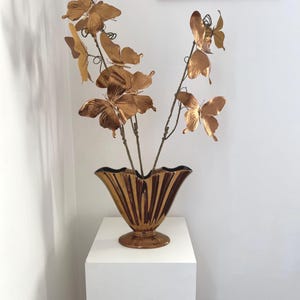 Puede incluir: Un arreglo decorativo con adornos de mariposas de color bronce sobre finos tallos, exhibido en un jarrón de bronce a juego. El jarrón tiene un diseño acanalado y se asienta sobre un pedestal blanco, creando una exhibición elegante.