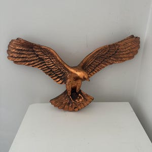 Könnte beinhalten: Eine bronzefarbene Adler-Skulptur mit ausgebreiteten Flügeln. Der Adler sitzt auf einem Sockel, mit detaillierten Federn auf den Flügeln und dem Körper. Die Skulptur wird auf einer weißen Oberfläche vor einer weißen Wand ausgestellt.