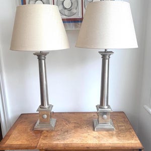 May include: A pair of table lamps featuring tall, silver-toned metal columns with square bases and circular decorative accents. Each lamp is topped with a tapered, off-white fabric shade, providing a balanced and symmetrical lighting arrangement.