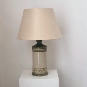 Könnte beinhalten: Eine Keramik-Tischlampe mit beigem Lampenschirm. Der Sockel ist zylindrisch, mit einem hellbeigen Korpus und dunkelgrünen Akzenten. Die Lampe steht auf einem weißen Sockel, der ihr Design zur Geltung bringt. Die Höhe der Lampe beträgt ca. 61 cm.