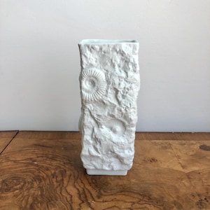 May include: White ceramic vase with a textured surface resembling rock. The vase has a square shape and features raised, embossed designs of fossilised ammonites.