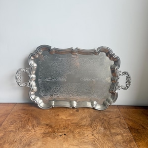 Puede incluir: Una bandeja de servicio rectangular de metal plateado con un borde festoneado y asas ornamentadas y detalladas en ambos lados. El centro de la bandeja está decorado con un intrincado patrón floral grabado y un espacio rectangular liso y vacío.