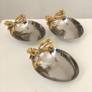May include: Three silver oval-shaped bowls with hammered metal finishes and gold floral accents. The bowls are ideal for serving snacks or small dishes.