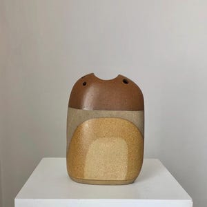 Puede incluir: Un jarrón de cerámica marrón y beige con forma rectangular redondeada y una abertura ancha. El jarrón tiene un diseño geométrico con una parte superior marrón, una sección media beige y una parte inferior amarilla.