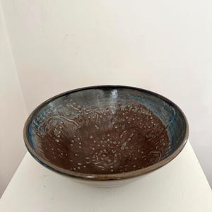 Op de afbeelding: Een handgemaakte keramische kom met een donkerbruin interieur en een blauwzwarte rand. De kom heeft een textuuroppervlak met kleine witte stippen en een glanzende afwerking. De kom wordt gepresenteerd op een wit oppervlak.