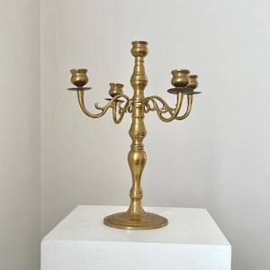 May include: A vintage brass candelabra with five arms. The candelabra has a central column with a round base and ornate, curved arms. The candle holders are round and slightly flared. The piece is displayed on a white surface.
