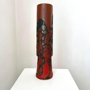 May include: A tall, cylindrical vase with a textured brown upper section and a flared, red base. The vase features a decorative pattern of black and silver shapes. The vase is displayed on a white surface.