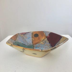 Może przedstawiać: Kwadratowa ceramiczna miska z kremowym zewnętrzem i kolorowym wnętrzem. Wnętrze ma wzór patchworkowy z odcieniami niebieskiego, pomarańczowego, czerwonego i zielonego. Małe czarne kółko znajduje się na środku miski.