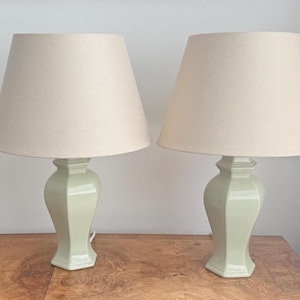 Może przedstawiać: Para lamp stołowych z jasnozielonymi ceramicznymi podstawami o fasetowanym, urnowatym kształcie. Każda lampa jest zwieńczona szerokim, zwężającym się kloszem bębnowym z neutralnej beżowej tkaniny, co zapewnia czysty i zrównoważony wygląd oświetlenia.
